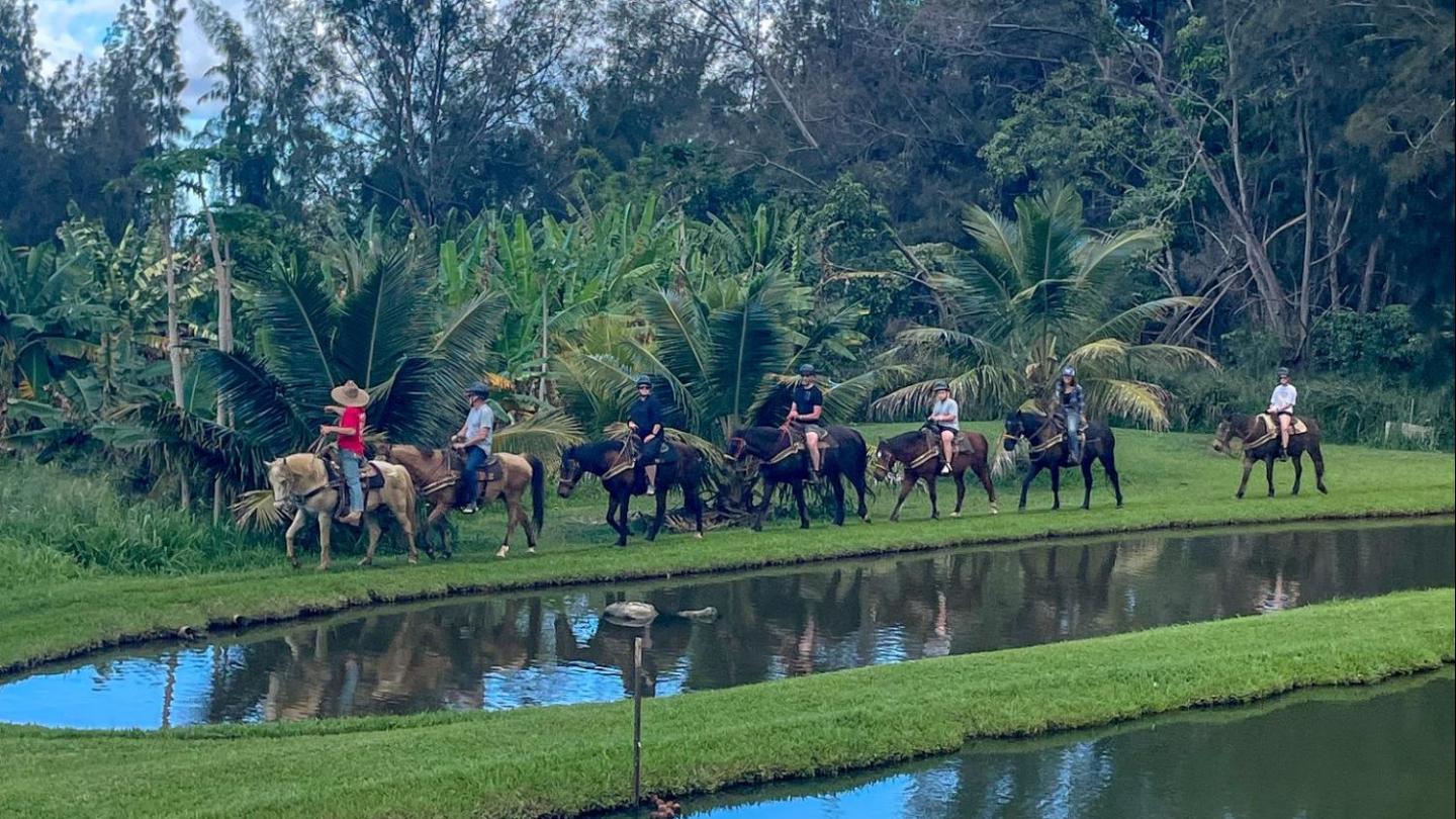 People riding horses beside a pond with trees in the background.