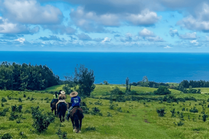 Three people horseback riding on a green hill overlooking the ocean.