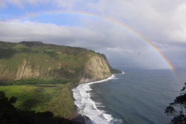 Coastal cliff with ocean waves and a rainbow in a cloudy sky.
