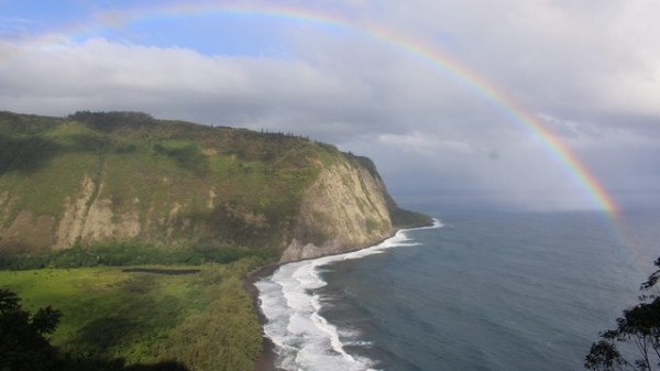 Coastal cliff with ocean waves and a rainbow in a cloudy sky.
